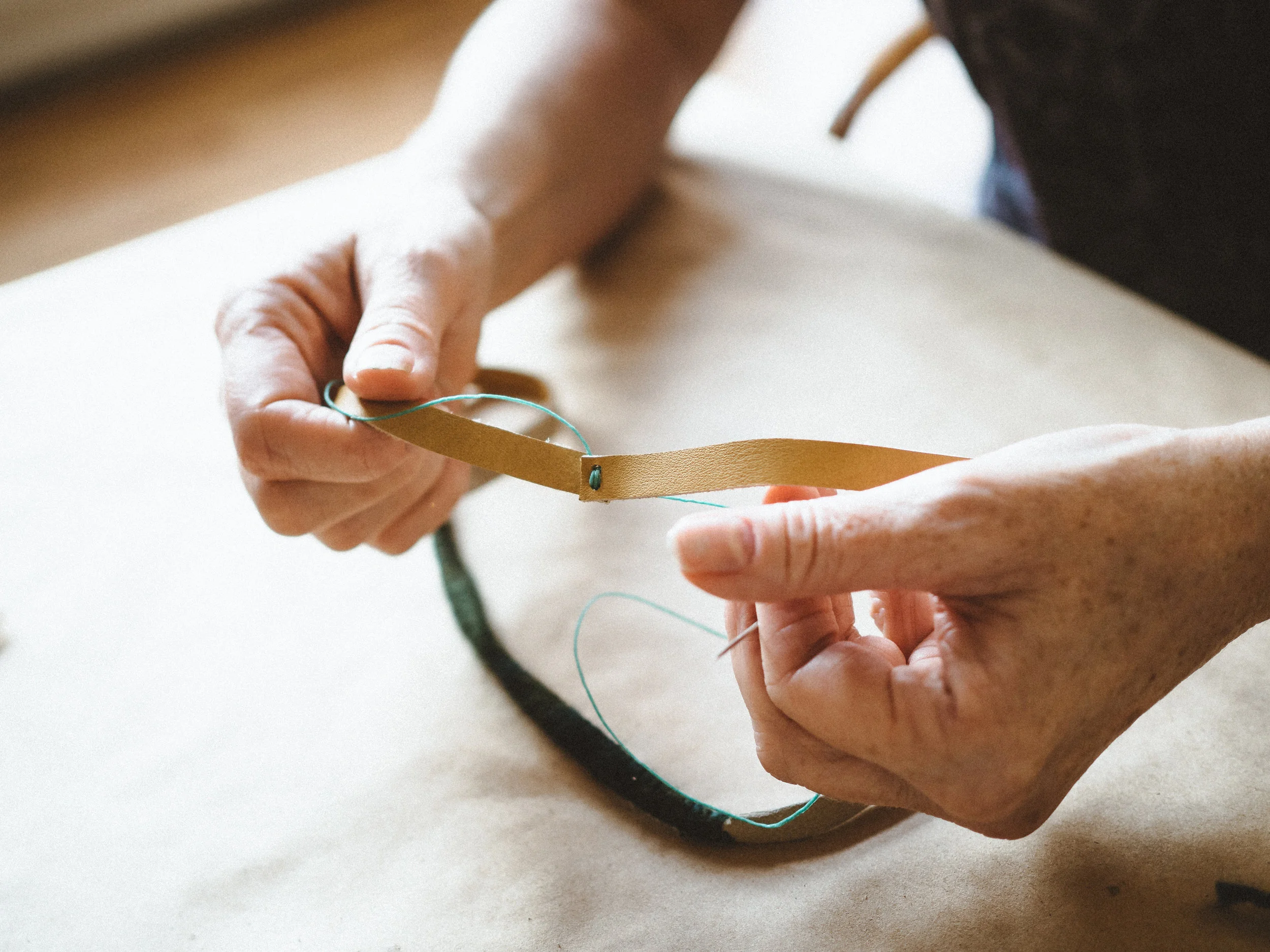 Close-up of two hands stitching a tan leather strip with teal thread and needle on a beige work surface, warm crafting mood